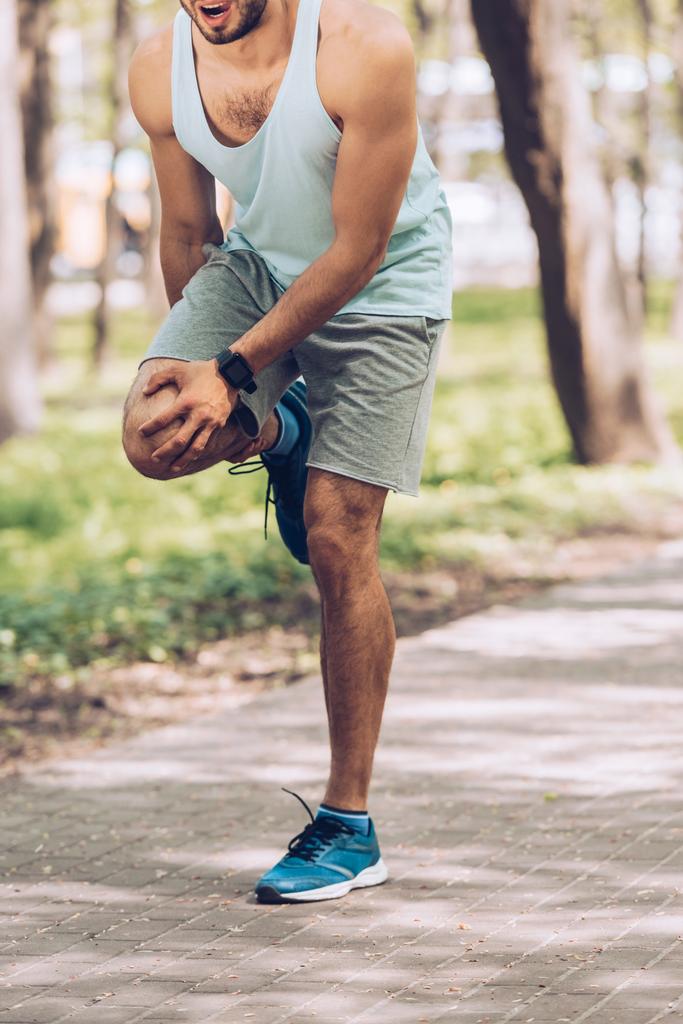 公園に立っている間に負傷した膝に触れるスポーツマンのトリミングショット - 写真・画像