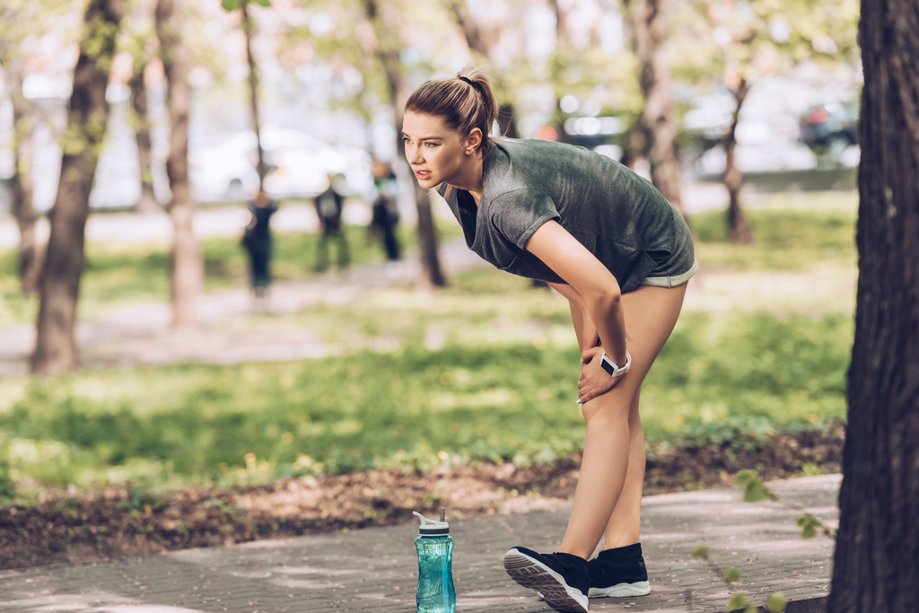 pretty sportswoman touching injured knee while standing in park near sport bottle - Photo, Image
