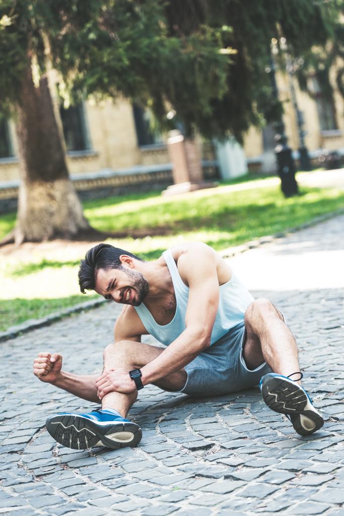 молодой спортсмен, страдающий от боли, сидя на тротуаре и касаясь поврежденной ноги
 - Фото, изображение