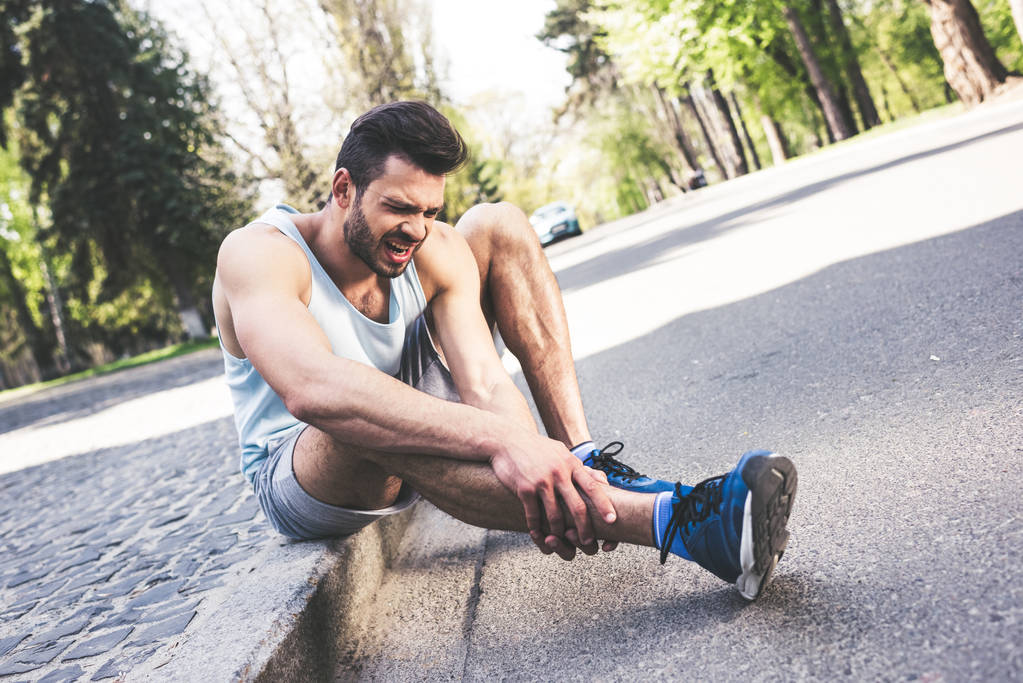 травмированный спортсмен, страдающий от боли, сидя на границе тротуара и касаясь ноги
 - Фото, изображение