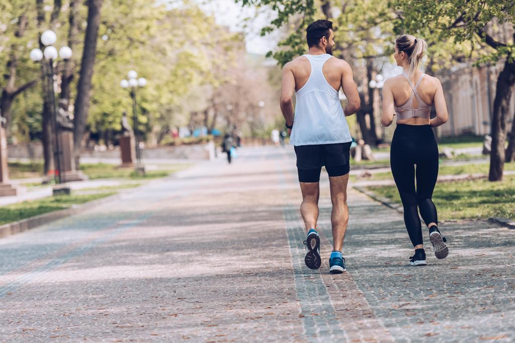 вид сзади на мужчину и женщину в спортивной одежде, бегущих вдоль аллеи парка
 - Фото, изображение