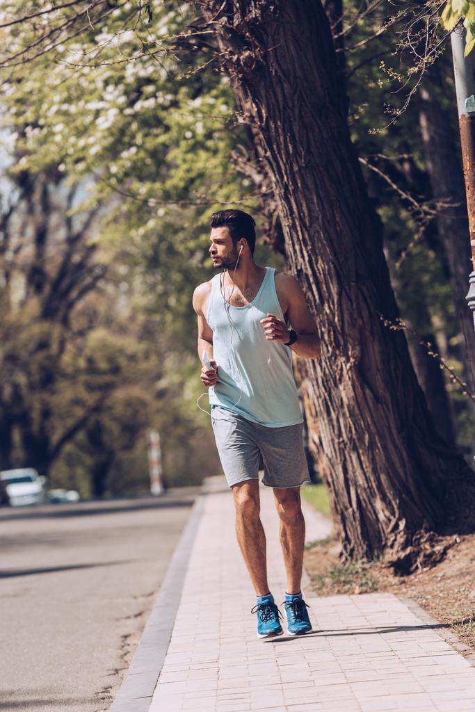 handsome sportsman listening music in earphones while jogging alone pavement near roadway - Photo, Image