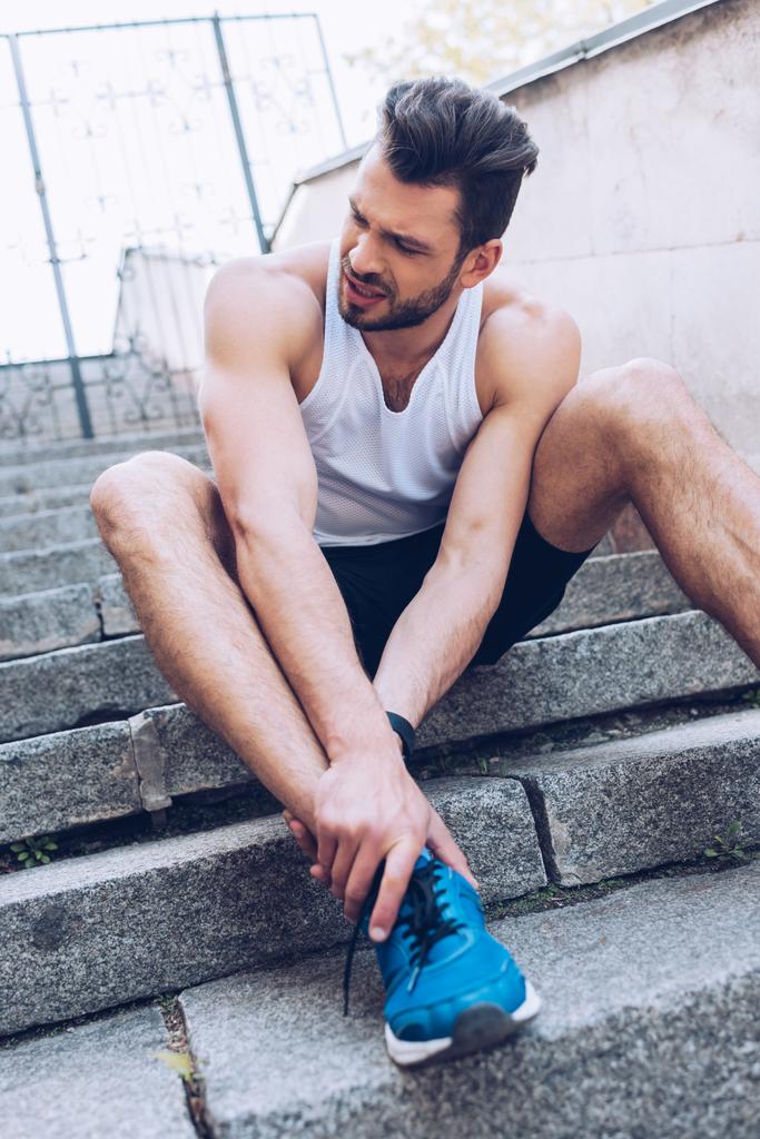 расстроенный спортсмен, сидящий на лестнице и страдающий от боли при прикосновении к травме колена
 - Фото, изображение