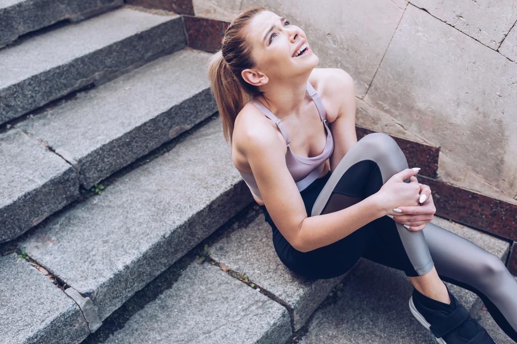 injured sportswoman looking up while sitting on stairs and suffering from pain in leg - Photo, Image