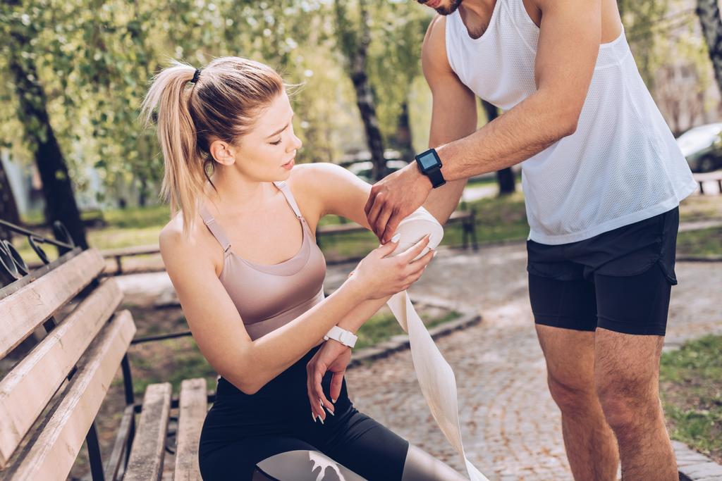 man in sportswear bandaging injured elbow of young sportswoman sitting on bench in park - Photo, Image