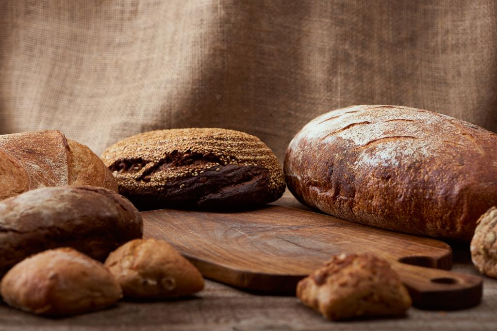 selective focus of delicious loaves of bread near wooden chopping board with sackcloth on background - Photo, Image