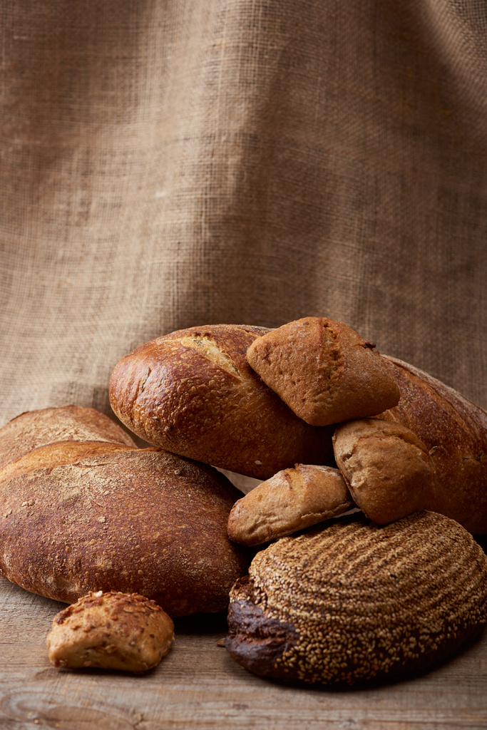 вкусные буханки хлеба и булочки на деревянном столе со вретищем на заднем плане
 - Фото, изображение