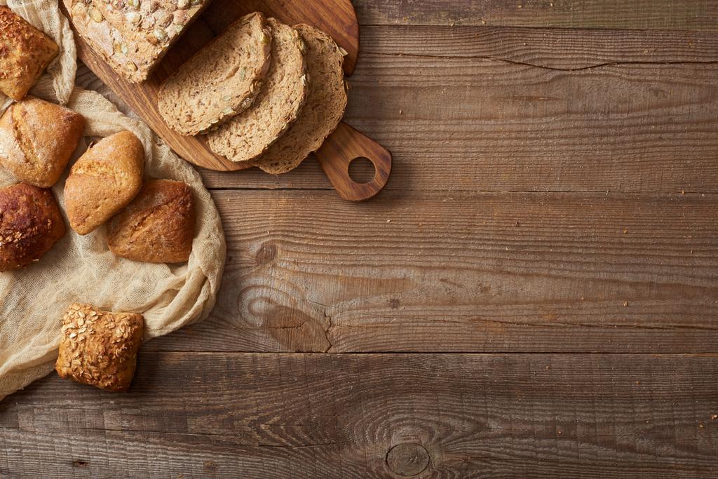 вид зверху на скибочки свіжого хліба на рубаній дошці та булочки на тканині на дерев'яному столі
 - Фото, зображення