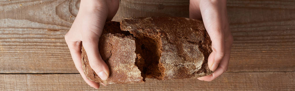 top view of woman holding fresh homemade brown bread above wooden table, panoramic shot - Photo, Image