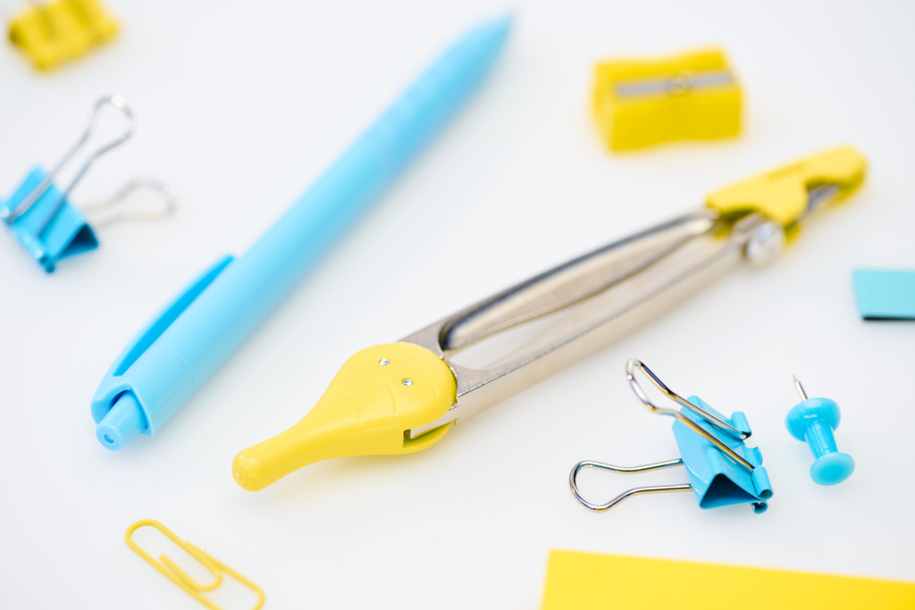селективный фокус желтой и синей канцелярских принадлежностей с скрепками, компасами, точилкой для карандашей и ручкой на белом фоне
 - Фото, изображение