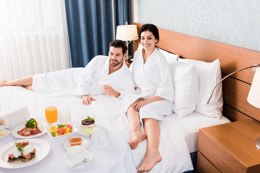 happy man and woman with barefoot looking at breakfast in hotel room  - Photo, Image