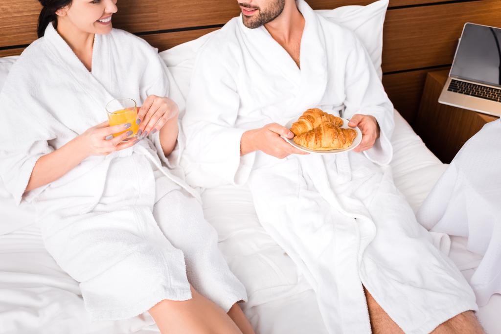 vue recadrée de l'homme joyeux tenant des croissants tout en étant couché près de la femme avec un verre de jus d'orange
  - Photo, image