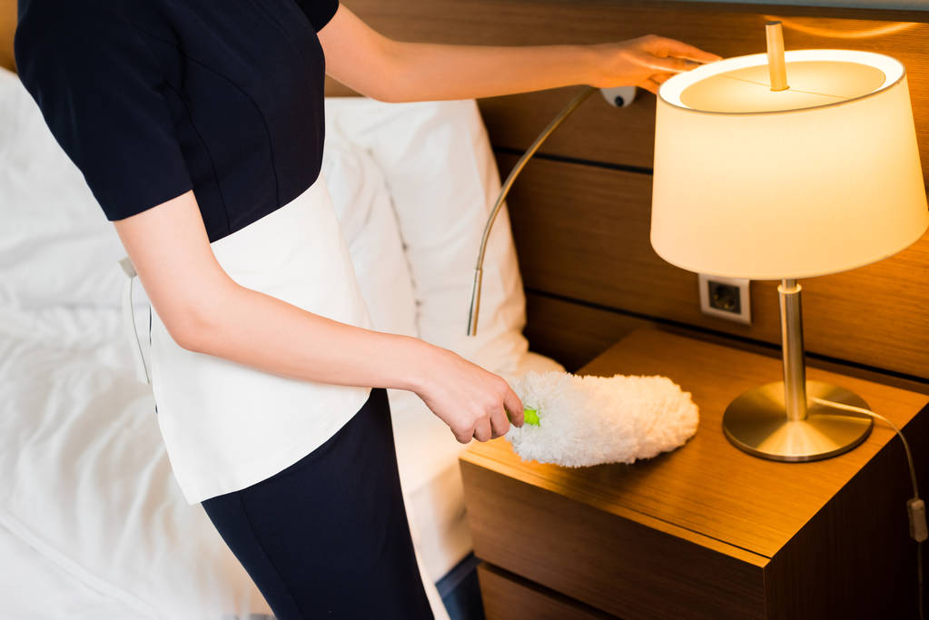 περικομμένη θέα της υπηρέτριας σε λευκή ποδιά κρατώντας ξεσκόνισμα και τον καθαρισμό δωμάτιο ξενοδοχείου  - Φωτογραφία, εικόνα