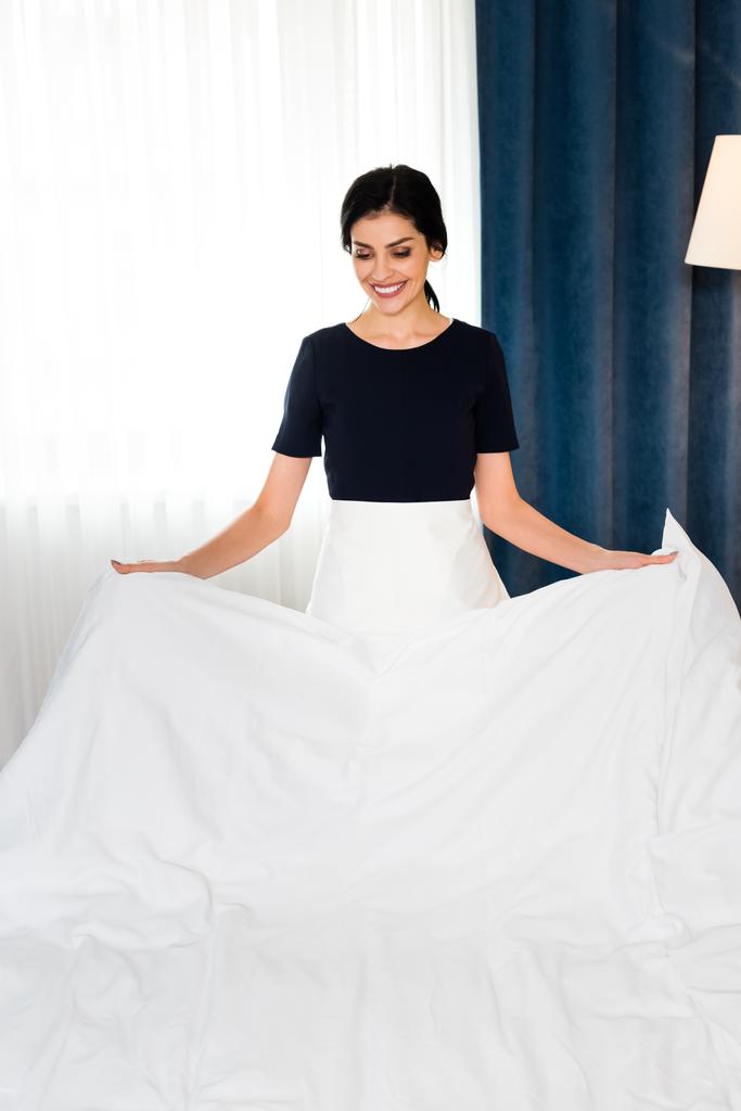 ευτυχισμένος υπηρέτρια κρατώντας λευκό φύλλο κρεβάτι στο δωμάτιο του ξενοδοχείου  - Φωτογραφία, εικόνα