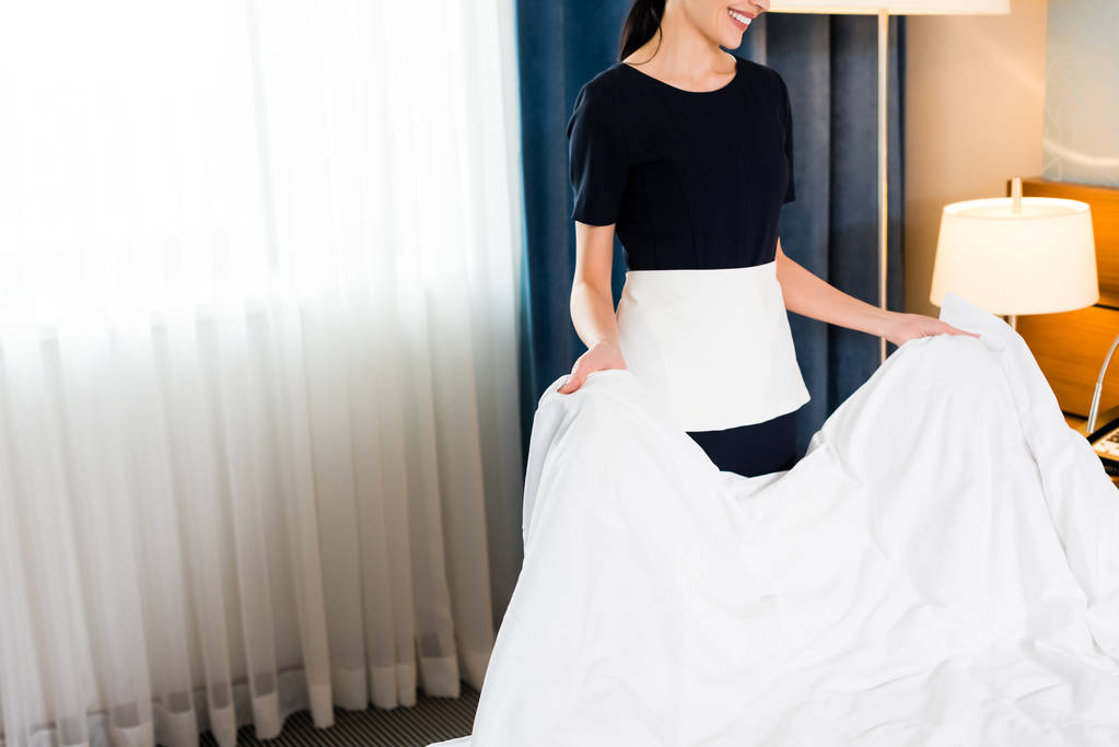 vue recadrée de gaie femme de ménage tenant drap de lit blanc dans la chambre d'hôtel
  - Photo, image