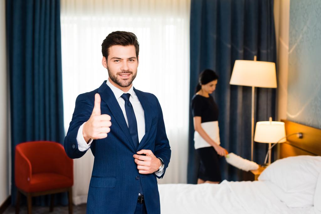 Selektiver Fokus der glücklichen Empfangsdame zeigt Daumen hoch neben Dienstmädchen im Hotelzimmer  - Foto, Bild
