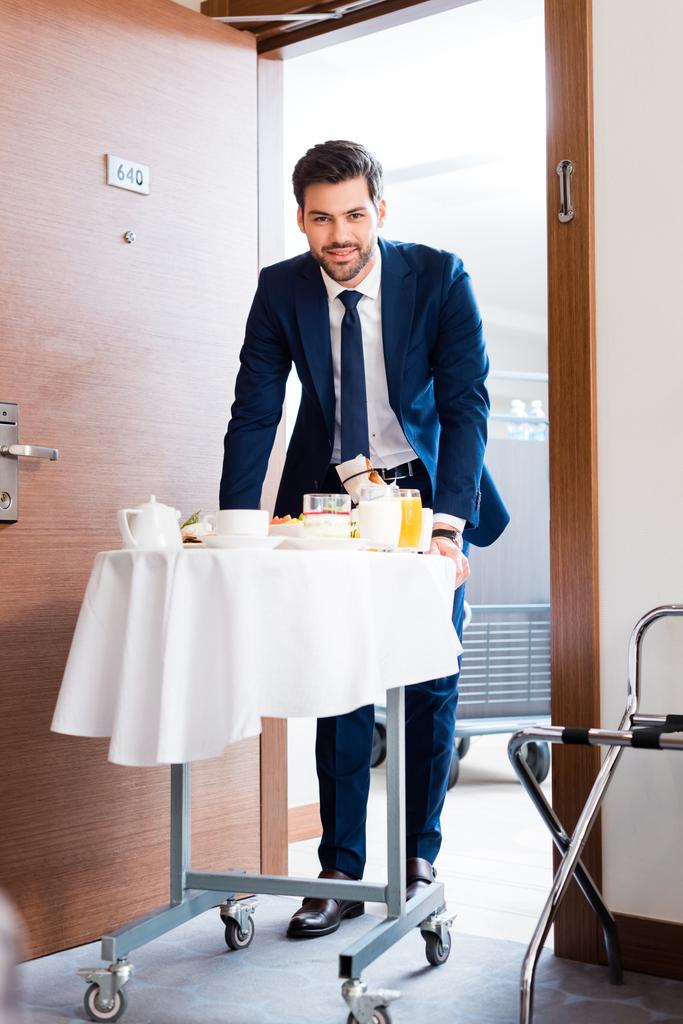 selektywne skupienie wesołej recepcjonistka przynosząc śniadanie na hotelowej wózek spożywczy  - Zdjęcie, obraz