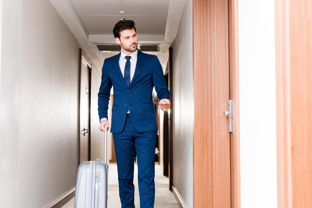όμορφος άνθρωπος στέκεται με αποσκευές και κατοχή κάρτα ξενοδοχείου στο διάδρομο του ξενοδοχείου  - Φωτογραφία, εικόνα