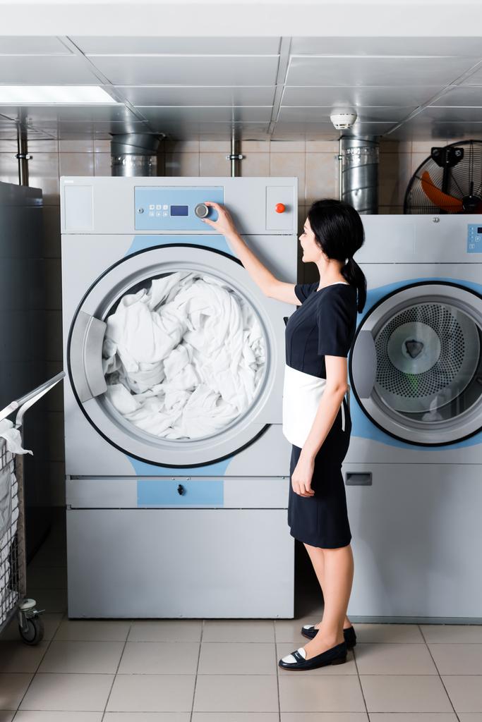 Горничная брюнетки стоя и касаясь кнопки на стиральной машине в прачечной
  - Фото, изображение