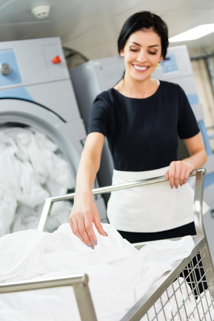selektivní zaměření šťastné služebné u koše s bílými prostěradly v prádelně  - Fotografie, Obrázek