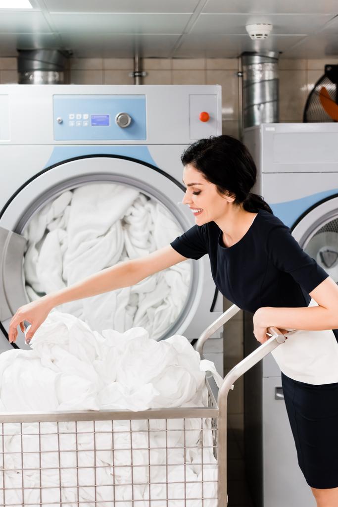 alegre mucama tocando sábanas mientras está de pie cerca de lavadoras en la lavandería
  - Foto, imagen