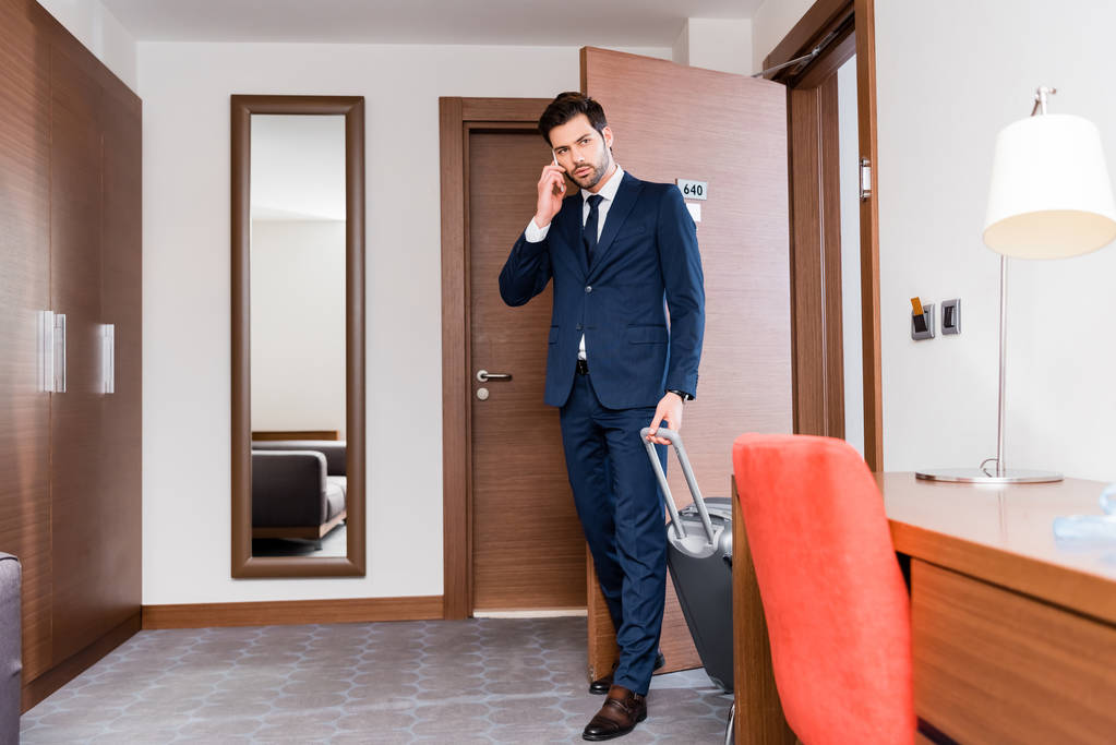 bel homme d'affaires parlant sur smartphone et debout avec valise dans la chambre d'hôtel
  - Photo, image