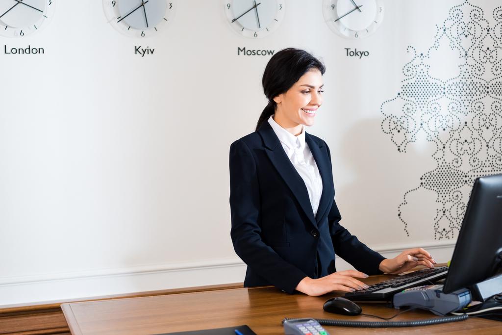 Gelukkig brunette receptioniste in formele slijtage staande in de buurt van computer monitor in Hotel  - Foto, afbeelding
