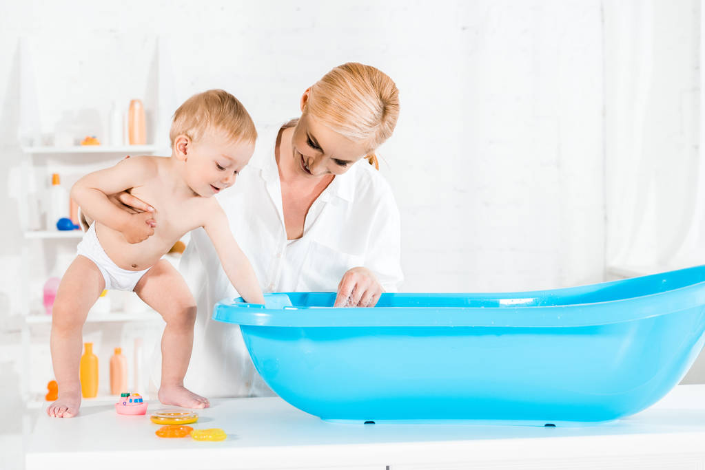 щаслива блондинка мати дивиться на дитячу ванну і тримає милий маленький син у ванній
  - Фото, зображення