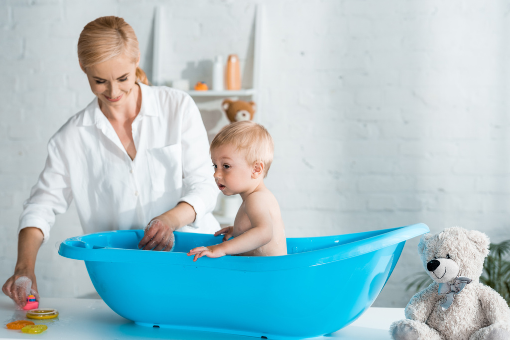 cheerful mother looking at cute toddler son taking bath at home  - Photo, Image