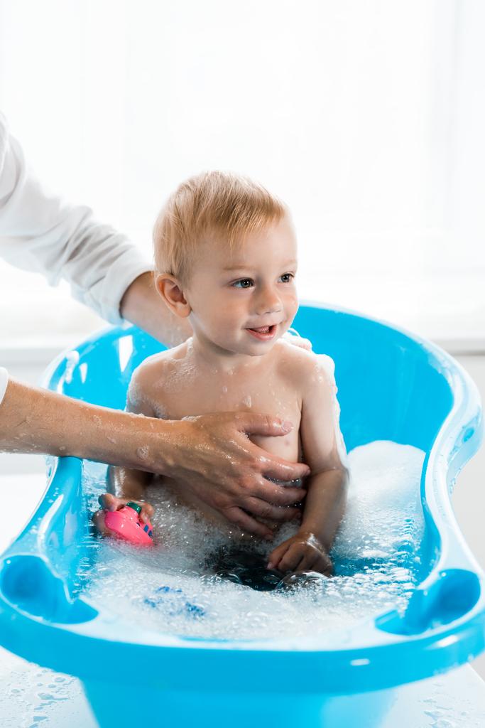 青い赤ちゃんのバスタブで幸せな幼児の子供を洗う母のトリミングビュー  - 写真・画像