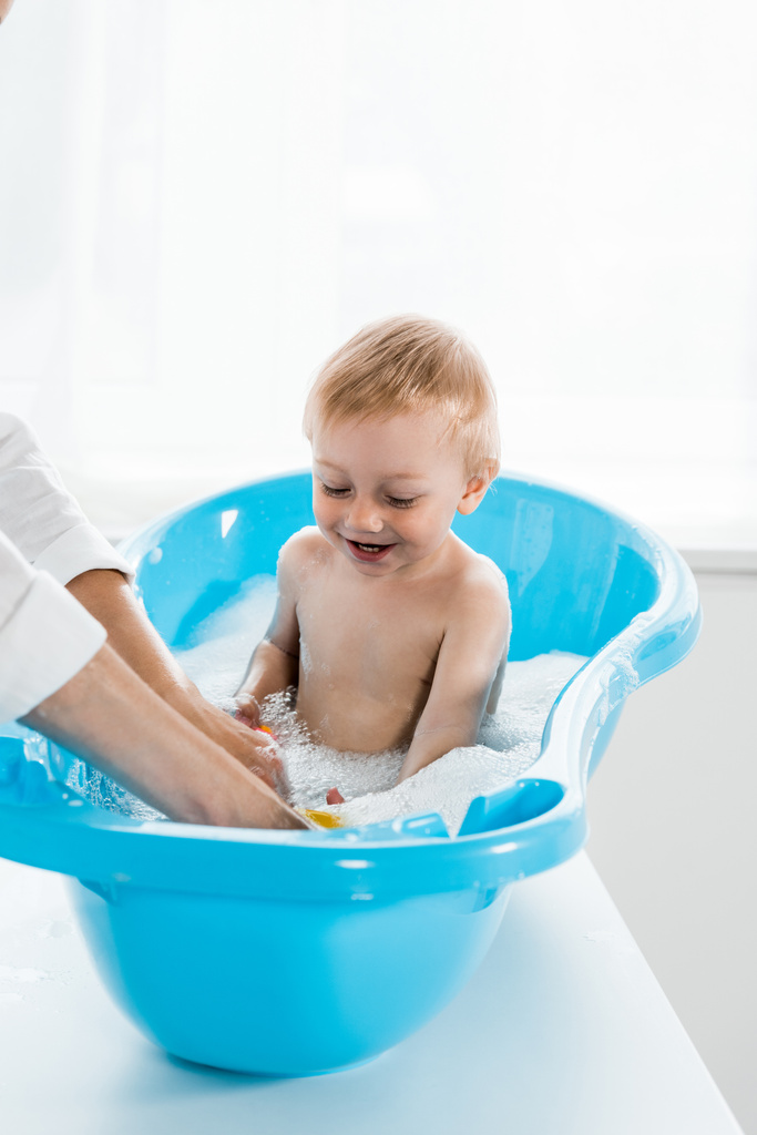 青い赤ちゃんのバスタブで笑顔の幼児の子供を洗う母親のトリミングビュー  - 写真・画像