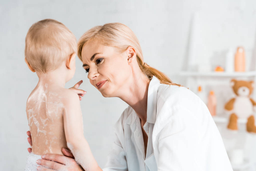 attraktive blonde Mutter schaut auf Kleinkind Sohn zeigt mit Finger im Badezimmer  - Foto, Bild