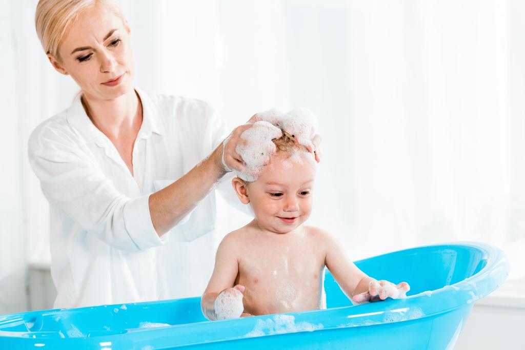 aantrekkelijke blonde moeder wassen hoofd van schattige peuter zoon in badkamer  - Foto, afbeelding