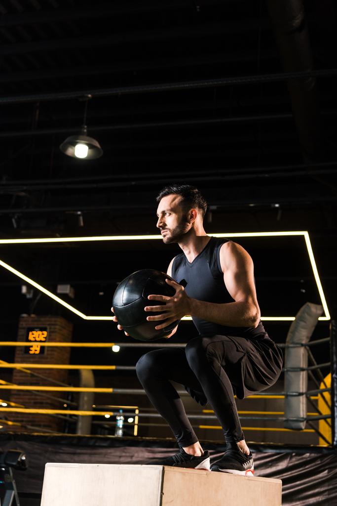 low angle view of handsome sportsman doing squat on squat box while holding ball - Photo, Image