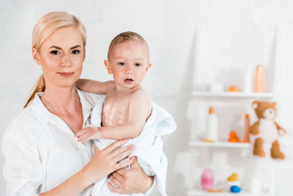 attractive blonde mother holding in arms wet toddler son in bathroom  - Photo, Image
