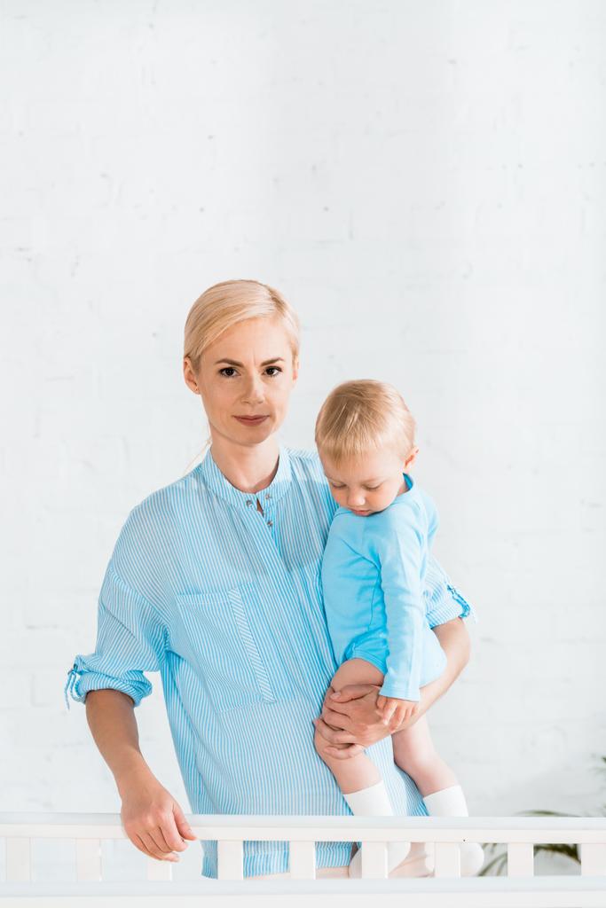 aantrekkelijke moederholding in Arms peuter zoon en staande in de buurt van baby wieg thuis - Foto, afbeelding