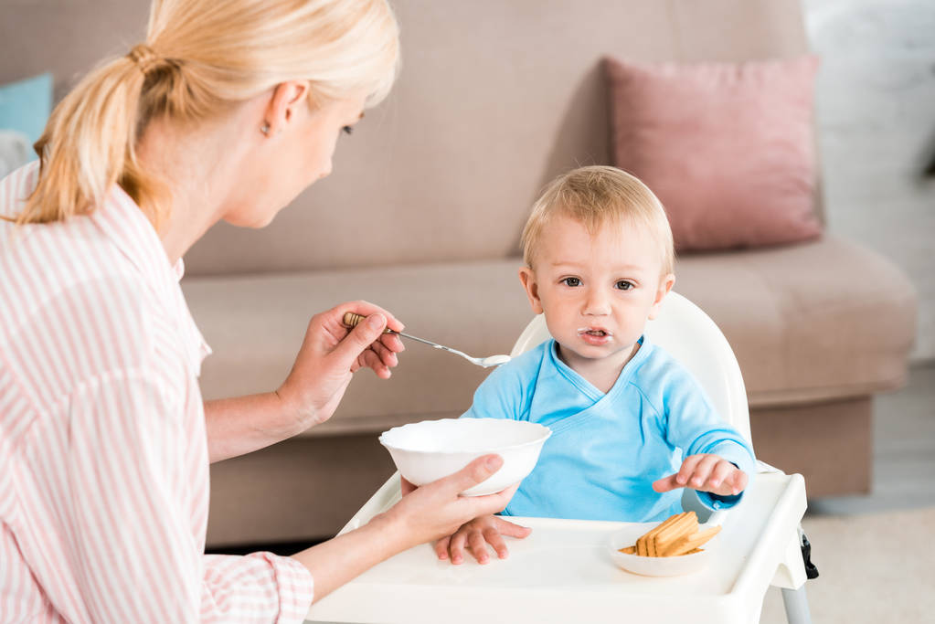 selective focus of blonde mother holding bowl and feeding cute toddler son at home  - Photo, Image