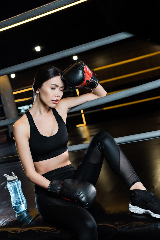 mujer en guantes de boxeo tocando la cabeza cerca de la botella de deporte en el gimnasio
  - Foto, Imagen