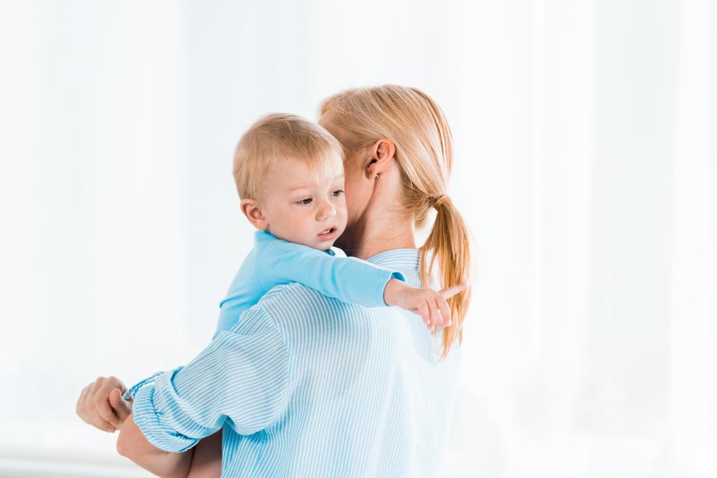 Rückansicht einer blonden Frau, die ihren niedlichen Kleinkindsohn im Arm hält und mit dem Finger auf ihr Zuhause zeigt  - Foto, Bild