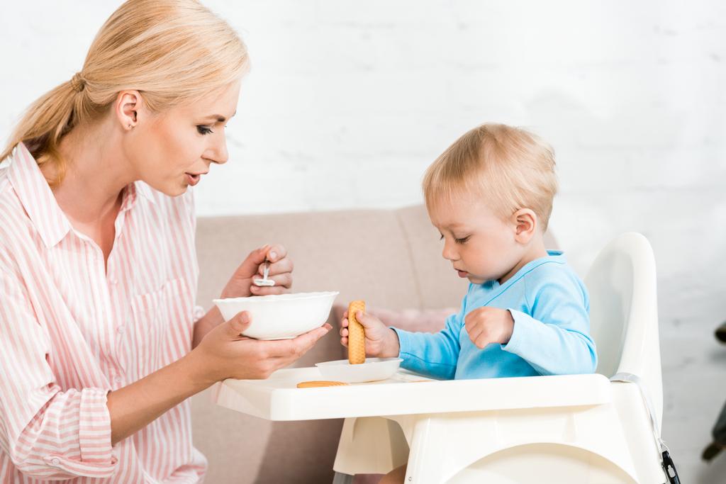 attractive mother holding spoon with baby food near cute toddler son sitting in feeding chair  - Photo, Image