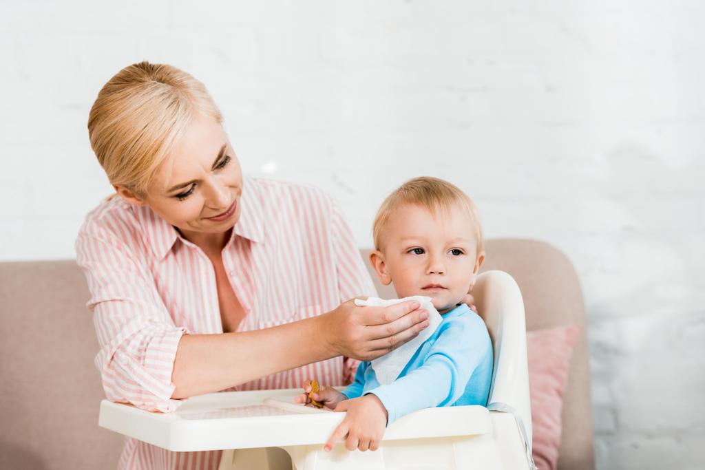 glückliche blonde Mutter hält Serviette in der Nähe von niedlichen Kleinkind Sohn sitzt in Fütterungsstuhl  - Foto, Bild