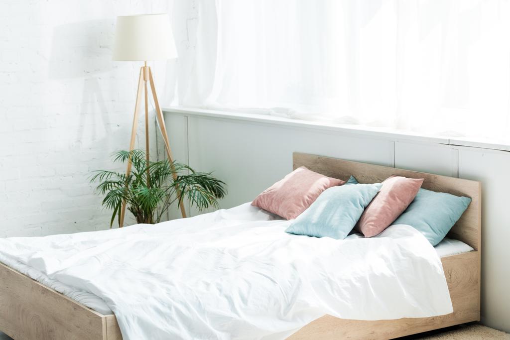 Bett mit weißer Bettwäsche, blauen und rosa Kissen in der Nähe von Stehlampe und Pflanze - Foto, Bild