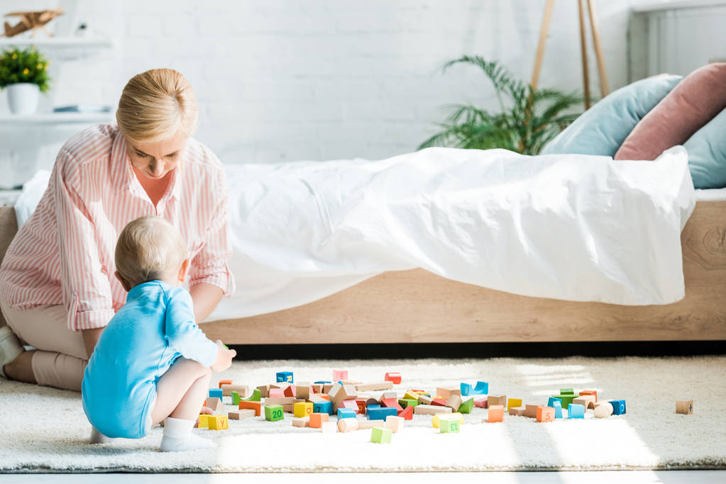 atraente loira mãe sentado perto de criança filho e brinquedos blocos no quarto
  - Foto, Imagem