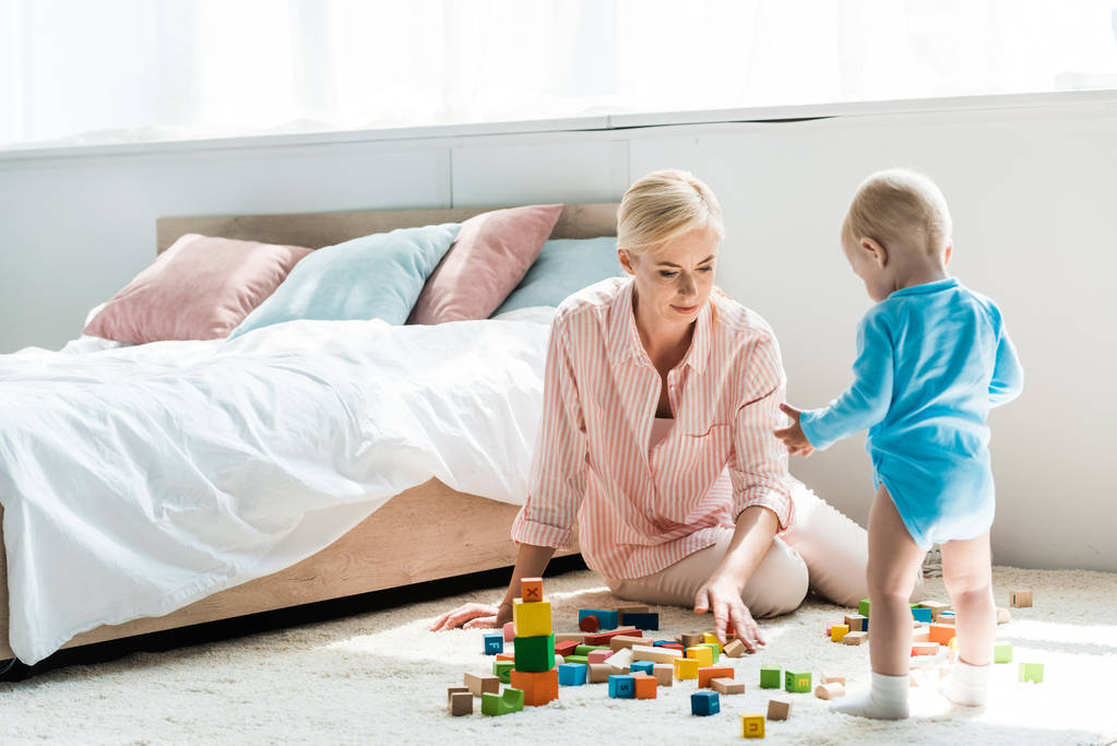 blonde moeder zittend op tapijt in de buurt van peuter zoon spelen met bouwstenen  - Foto, afbeelding