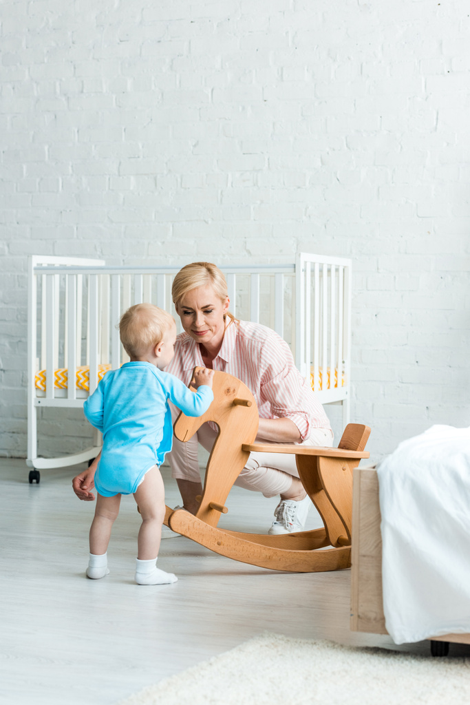 cute toddler son standing near attractive blonde mother and wooden rocking horse  - Photo, Image