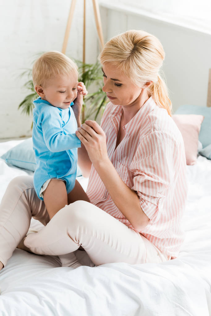 aantrekkelijke blonde moeder houden handen met peuter zoon terwijl zittend op bed  - Foto, afbeelding