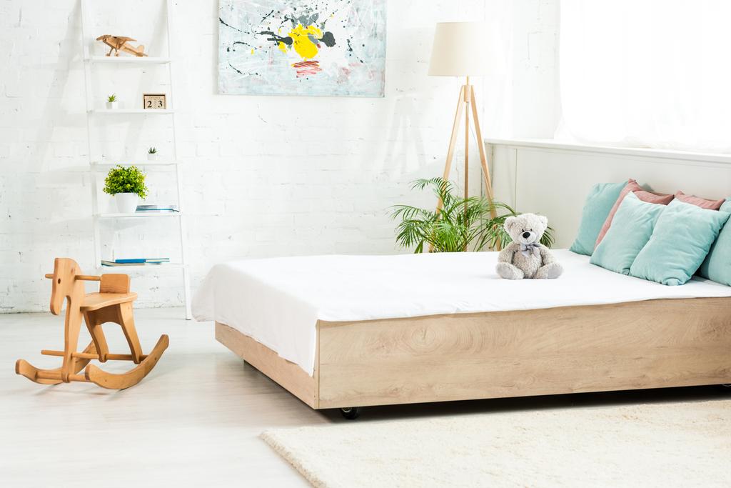 cama com roupa de cama branca perto de ursinho de pelúcia e cavalo de balanço de madeira no quarto
  - Foto, Imagem