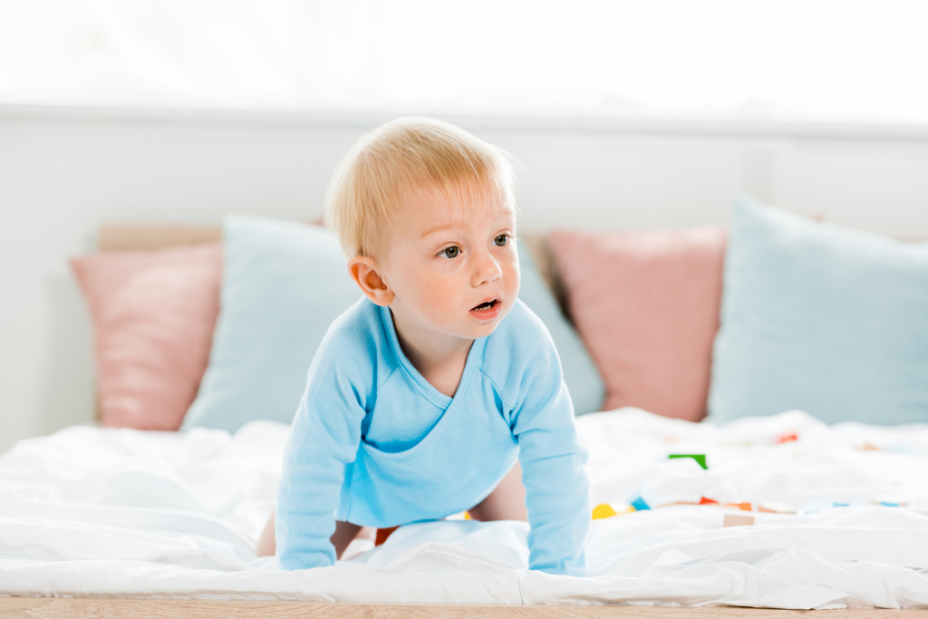 Überraschtes Kleinkind krabbelt mit weißer Bettwäsche zu Hause auf Bett  - Foto, Bild