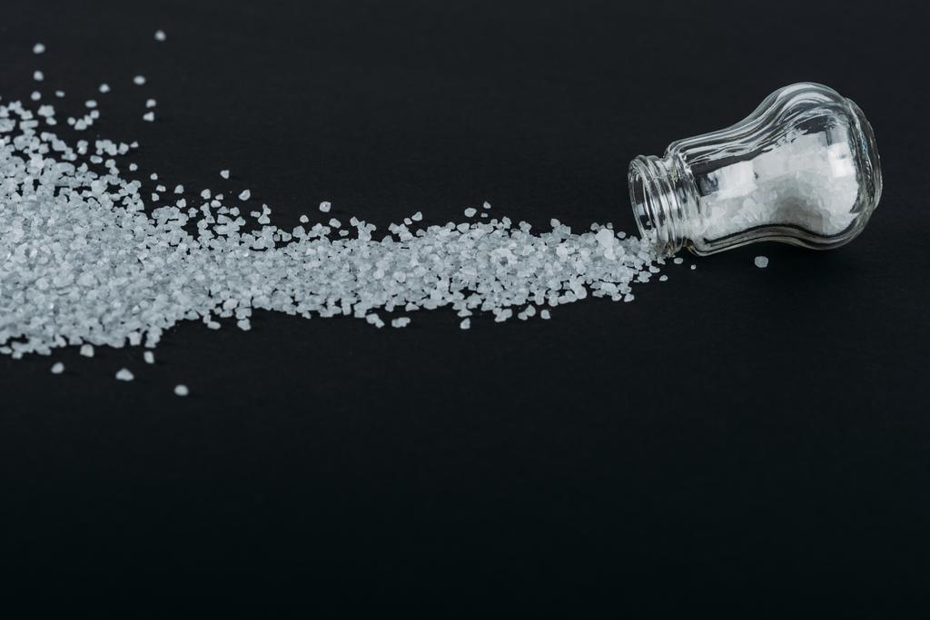 top view of salt shaker near sprinkled salt crystals on black background - Photo, Image