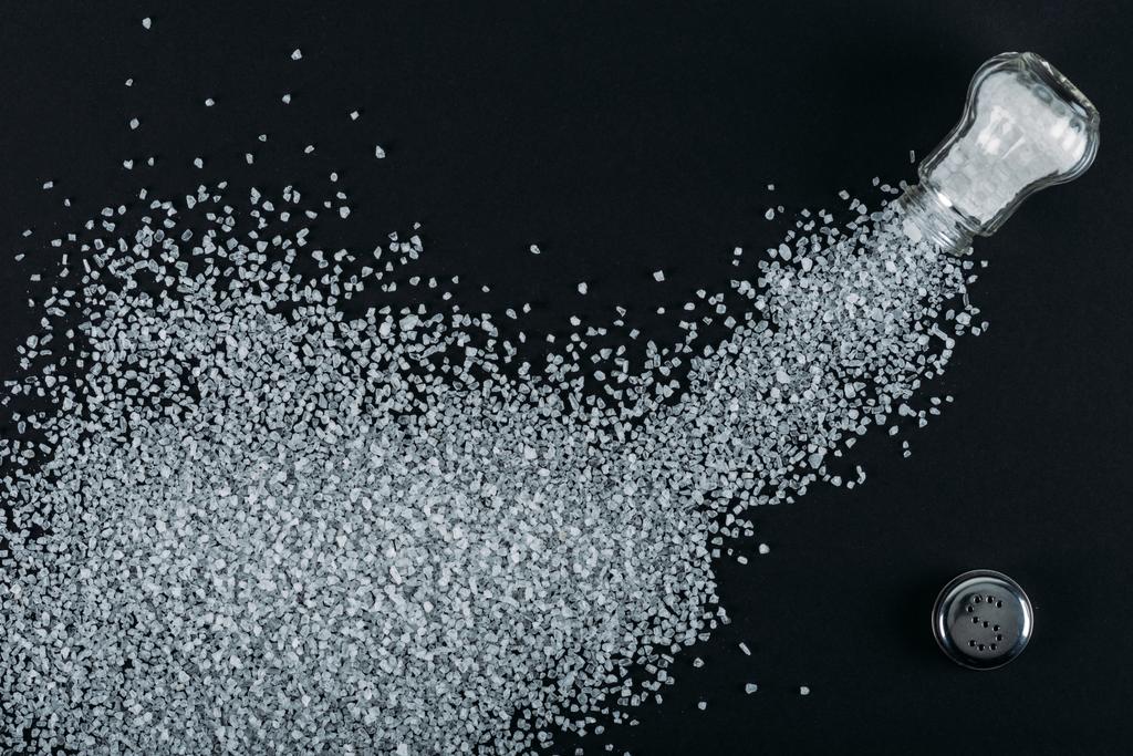 Top uitzicht van zout kristallen besprooit in de buurt van zout shaker op zwarte achtergrond - Foto, afbeelding